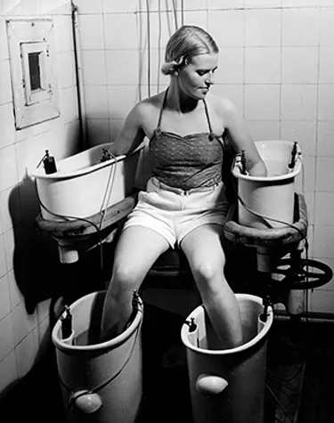 A young woman holds her arms and legs in four water baths with electric current circa 1938.
